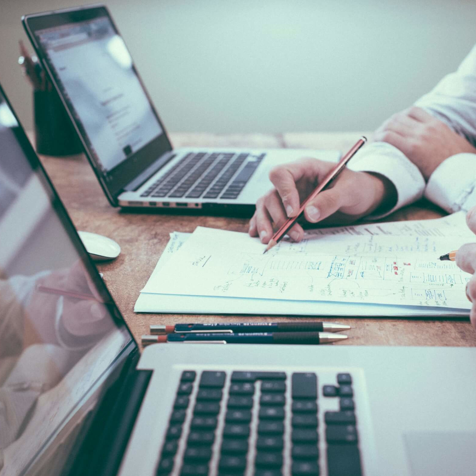 Personne analysant des documents et prenant des notes sur un bureau avec deux ordinateurs portables ouverts.